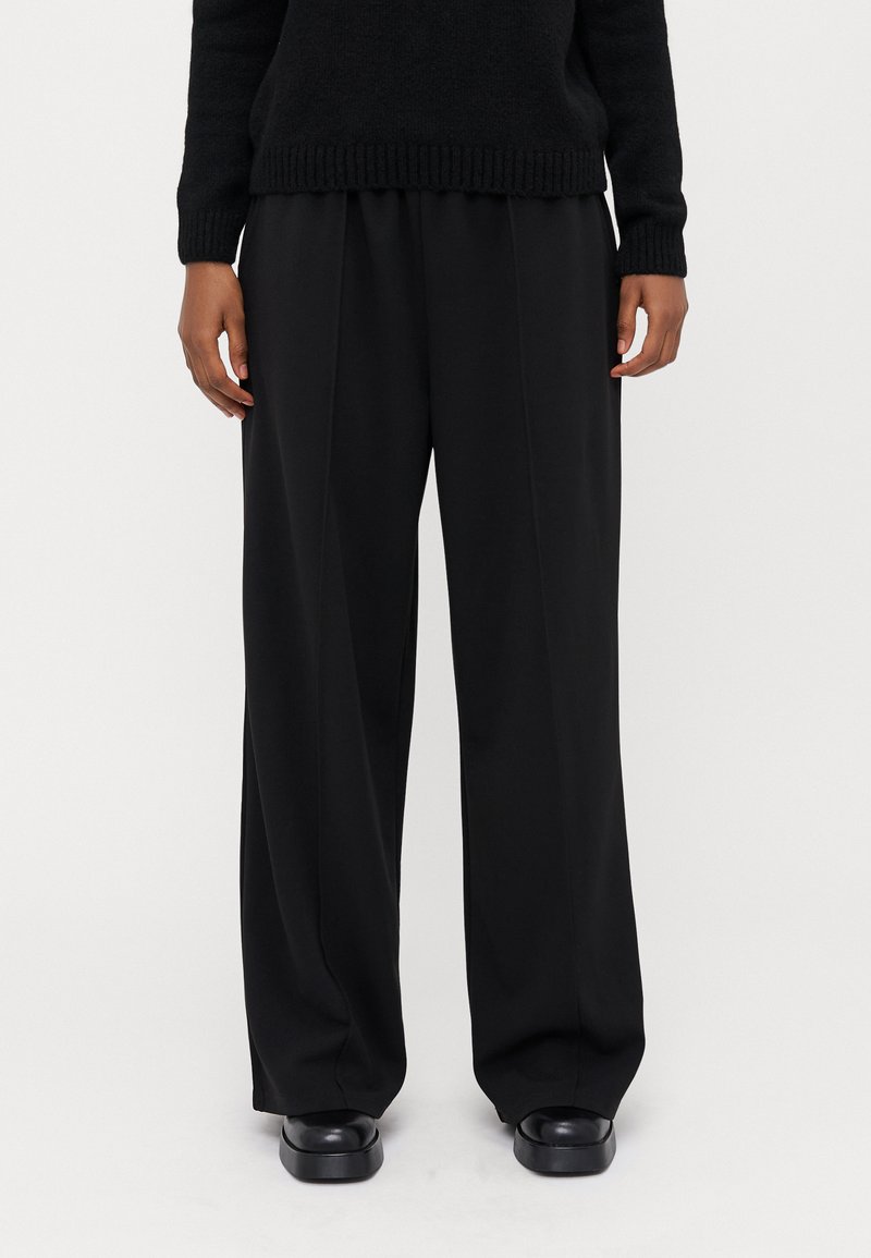 Pantalons larges noirs en tissu lisse, avec une taille haute et un devant plissé. Associés à des chaussures à plateforme noires.