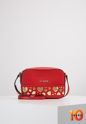 Sac bandoulière rouge avec découpes en forme de cœur dorées et logo Love Moschino, présenté sur une surface blanche avec un fond uni.