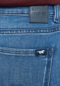 Poche en denim bleu sur un jean avec un patch en cuir de la marque Mustang et une petite étiquette arborant un logo de cheval blanc en train de galoper.