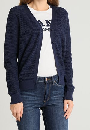 Frau trägt eine marineblaue, strukturierte, knöpfbare Strickjacke über einem weißen T-Shirt mit Grafik und blauen Jeans, steht vor einem einfarbigen Hintergrund.