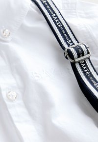 White cotton shirt featuring embroidered "BAKER" text, complemented by a navy and white woven strap with silver hardware. Button detail visible.