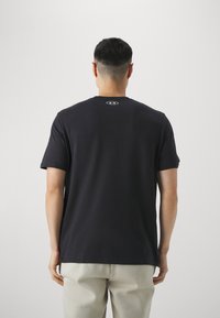 Muž v černém tričku s krátkým rukávem a malým bílým logem na horní části zad, oblečený do světlých kalhot, stojící před jednobarevným pozadím.