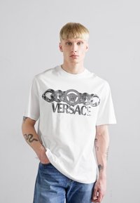 Valge puuvillane T-särk, millel on musta ketimustriga disain ja ümmargune logo, kus on tugevasti väljaprinditud "VERSACE" tekst rinnal.