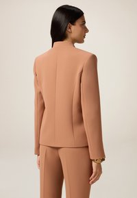 Blazer aderente color pesco con colletto alto, spalle strutturate e maniche lunghe, abbinato a pantaloni coordinati. Tessuto dalla texture liscia.