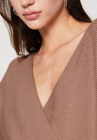 Top in maglia a coste marrone con scollatura a V profonda, caratterizzato da una texture morbida e una silhouette aderente, presentato su uno sfondo neutro.