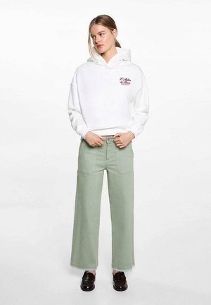 Bijela hoodie s logom sprijeda i oversized krojem, uparen s hlačama širokih nogavica svijetlozelene boje s džepovima na zakrpu i strganym rubovima. Crne cipele.