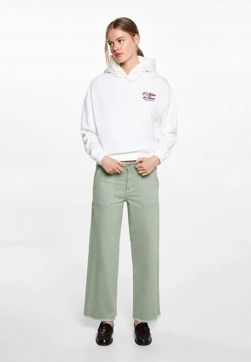 Bijela hoodie s logom sprijeda i oversized krojem, uparen s hlačama širokih nogavica svijetlozelene boje s džepovima na zakrpu i strganym rubovima. Crne cipele.