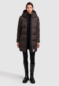 Donkerbruine pufferjas met capuchon, voorzien van een volledige rits, zijzakken en een logo-accent, gecombineerd met zwarte leggings en kniehoge laarzen.
