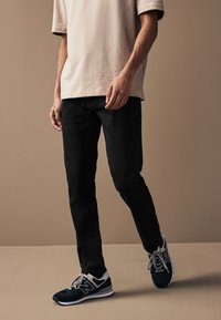 Schwarze Hose mit schmaler Passform; kombiniert mit einem beigefarbenen T-Shirt mit Rundhalsausschnitt und marineblauen Sneakern mit grauen Akzenten und weißen Sohlen.