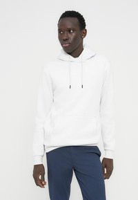 Άνδρας με σκουρόχρωμο δέρμα που φορά άσπρη κουκούλα και μπλε παντελόνι, στέκεται μπροστά σε απλό φωτεινό φόντο, κοιτάζοντας ελαφρώς προς το πλάι.