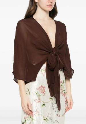 Donna che indossa uno scialle marrone trasparente legato davanti sopra una gonna bianca a fiori, mostrando una scollatura e l'addome scoperti.