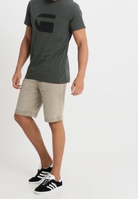 Czarnoszary T-shirt z krótkim rękawem z czarnym wzorem graficznym, zestawiony z beżowymi szortami i czarnymi butami w białe paski.