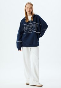 Sudadera azul marino de corte holgado con texto gráfico blanco, cuello con media cremallera y costuras blancas en contraste, combinada con pantalones anchos blancos.