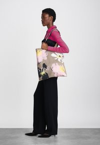 Blommönstrad toteväska i beige kanvas med en vit logotyp. Har stora svarta, rosa och gula blommor. Axelrem och rymlig design.