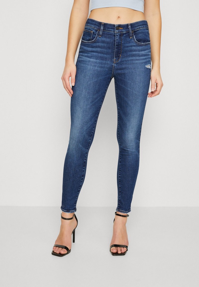 Jean skinny en denim délavé foncé avec une taille haute, présentant un léger effet froissé et un genou usé. Associé à des sandales noires à talons hauts.