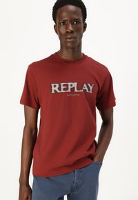Camiseta de algodón roja con escote redondo, mangas cortas y un logo gris "REPLAY" en el pecho. Ajuste casual con un mínimo de detalles.