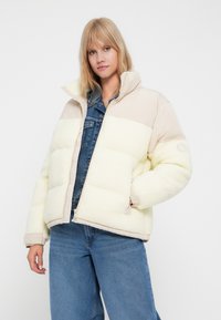 Geel en beige pufferjack met een fleece textuur, ritssluiting en elastische manchetten, gecombineerd met denim jeans.
