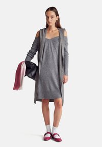 Šedé pletené šaty s žebrovaným lemem, doplněné dlouhým šedým cardiganem. Modelka drží černou kabelku a má na sobě vínové boty a bílé ponožky.