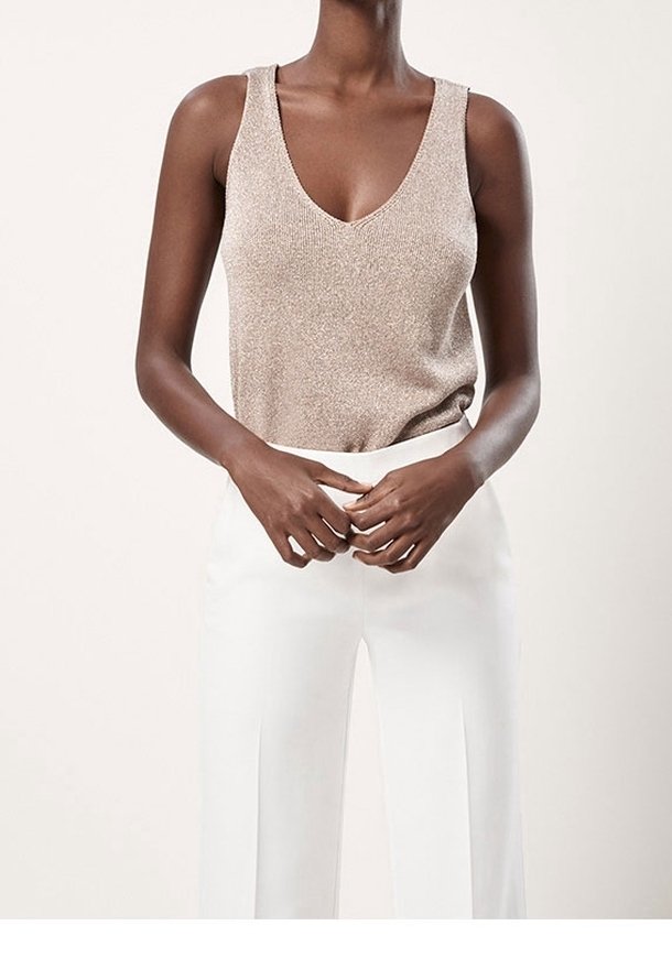 Personne portant un haut sans manches en maille beige avec un col en V et un pantalon blanc taille haute à jambes larges, debout, mains jointes.
