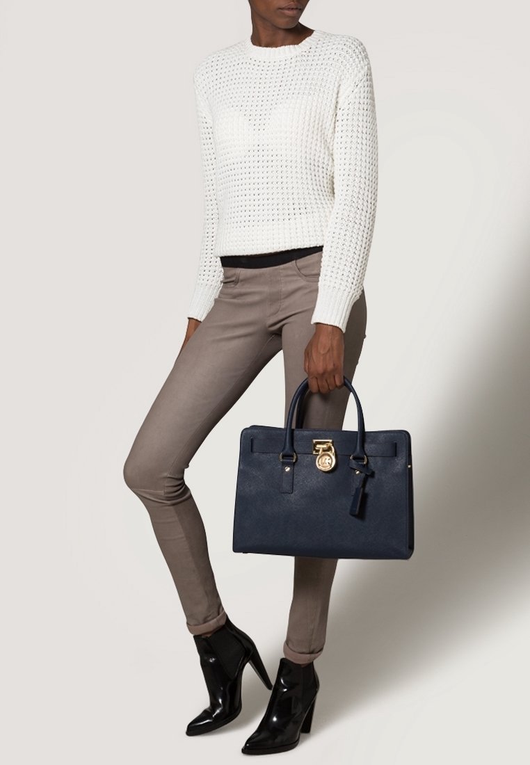 Femme portant un pull en maille blanc, un pantalon slim taupe, des bottines noires, tenant un sac à main bleu marine avec des ferrures dorées à ses côtés.