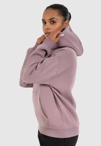 Sweatshirt à capuche violet clair avec une poche kangourou, des poignets côtelés et une texture douce, porté par un modèle se tenant de profil contre un arrière-plan neutre.