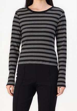 Kobieta ubrana w czarną bluzkę z długim rękawem z białymi poziomymi paskami i gładkie czarne spodnie, stojąca na jednolitym tle.