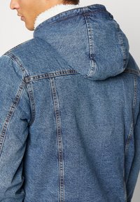 Jeansjacke mit Kapuze, in hellem Blau, mit strukturiertem Stoff und weißem Fleece-Futter am Kragen. Sichtbare Nähte.