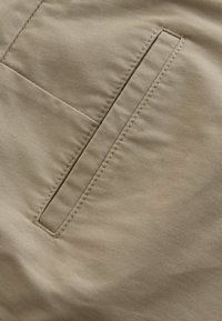 Béžová látka s hladkou textúrou, obsahujúca bočné vrecko s čistým prešívaním a obdĺžnikovým otvorom, typickým pre neformálne nohavice.