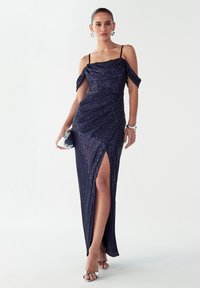 Večerna obleka temno modre barve z bleščicami, z rokavi, ki segajo čez ramena, stranskim razporkom in nabrano oprsje. Srebrne sandale z vrvicami in torbica dopolnjujejo videz.