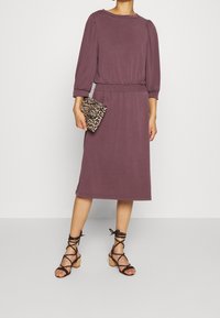 Lila knälång klänning med ballongärmar, elastisk midja, matchad med en leopardmönstrad kuvertväska och bruna sandaletter med klack.