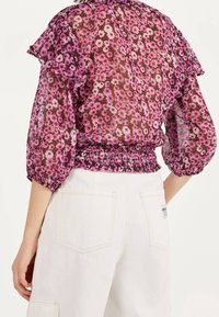 Kobieta w przezroczystej, różowej bluzce w kwiaty z falbanistymi rękawami oraz białych spodniach z wysokim stanem, pokazana tyłem.