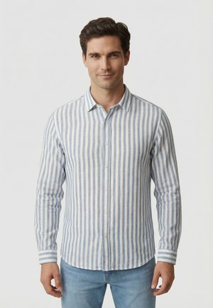 Hombre con camisa de botones de rayas verticales en azul claro y blanco de manga larga y vaqueros azul claro, de pie frente a un fondo blanco.