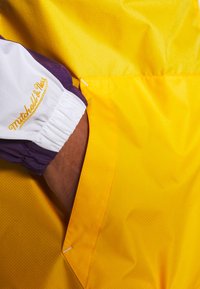 Gul jakke med lilla accenter; har en tekstureret finish, elastiske manchetter og et broderet "Mitchell & Ness" logo i guld på ærmet.