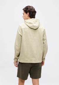 Άνδρας που φοράει μπεζ μπουφάν με κουκούλα και σκουροπράσινα σορτς, στέκεται γυρισμένος με την πλάτη σε ένα απλό φωτεινό φόντο.