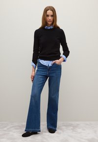 Suéter negro superpuesto a una camisa de cuello azul claro, combinado con jeans acampanados azules; el modelo se encuentra de pie contra un fondo neutro.