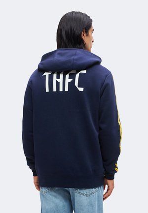 Muž s tmavými vlasmi po plecia, ktorý má na sebe tmavomodrú mikinu s kapucňou s "THFC" na chrbte a svetlomodré džínsy, stojí z profilu.