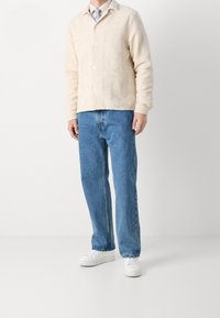 Ljust beige texturerad jacka med krage, knappstängning; matchad med blå löst sittande jeans och vita sneakers.