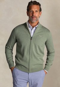 Groene ritstrui van zacht materiaal, met een hoge kraag en geribde boorden, gelaagd over een licht geruit overhemd.