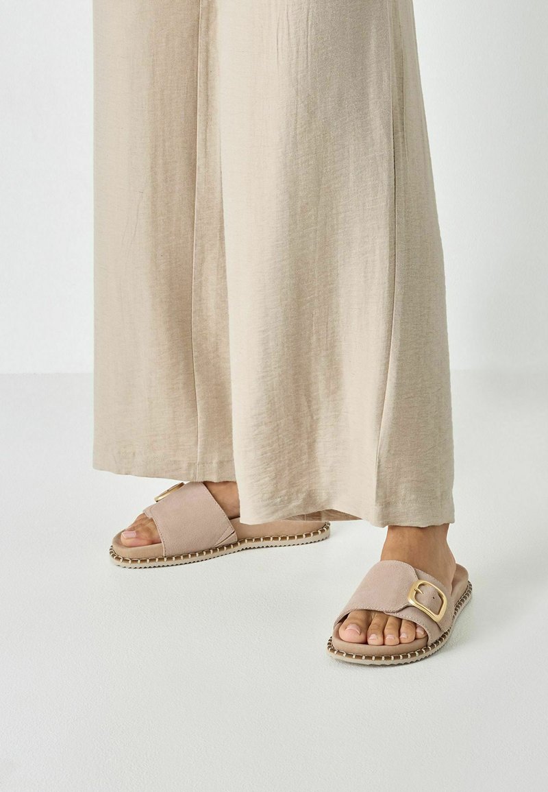 Sandales à glissière rose clair avec une bride texturée, un accent de boucle dorée et un détail de bord tissé, assorties à un pantalon beige ample.