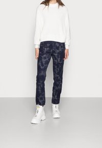 Biały sweter dziany, ciemnoniebieskie spodnie tie-dye i białe masywne sneakersy. Ubranie charakteryzuje się luźnym krojem i teksturowanym materiałem.
