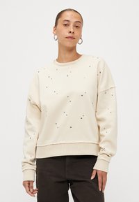 Κρέμα φούτερ με στρογγυλές στρας, στρογγυλή λαιμόκοψη, χαλαρή γραμμή και ριμπ μανσέτες. Συνδυάζεται με σκούρα παντελόνια.