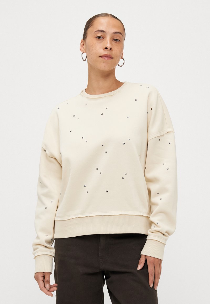 Κρέμα φούτερ με στρογγυλές στρας, στρογγυλή λαιμόκοψη, χαλαρή γραμμή και ριμπ μανσέτες. Συνδυάζεται με σκούρα παντελόνια.