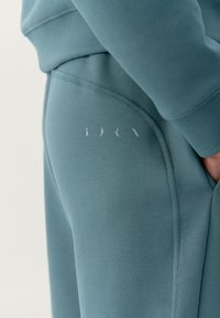 Tyrkysový sveter s hladkou textúrou, rebrovanými manžetami a diskrétnym bielym brandingom na boku, doplnený detailom švu.