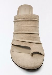 Bež semiš sandale z odprtim prstom, mehkim nabranim zgornjim delom in zanko za prste na ravnem podplatu na belem ozadju.