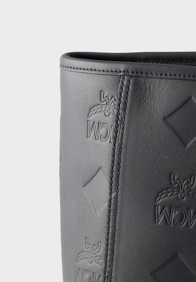 Gros plan sur le détail d'un sac en cuir noir avec des formes losanges embossées et le logo de la marque, coutures visibles le long du bord.