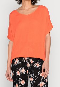 Kurzärmliges, V-Ausschnitt-Oberteil in leuchtendem Orange, mit lockerer Passform und weicher Textur. Kombiniert mit schwarzer Hose im Blumenmuster.