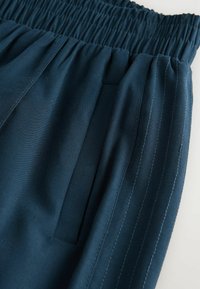 Elastische tailleband op donkerblauwe broek met een zijzak en verticale gestikte strepen langs de buitennaad.