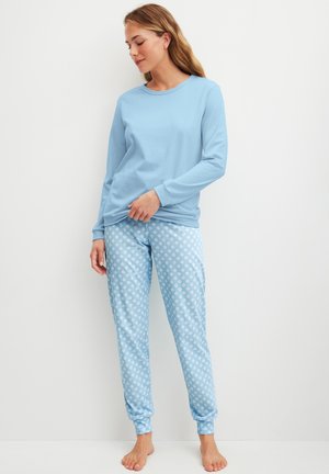Donna in piedi a piedi nudi che indossa una maglia a maniche lunghe azzurro chiaro e pantaloni da pigiama blu a pois, su sfondo bianco neutro.