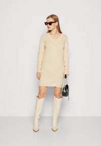 Šviesiai ruda megzta suknelė su V formos iškirpte ir ilgomis rankovėmis. Suderinta su blizgiais baltais batais iki kelių ir mažąja juoda rankine.