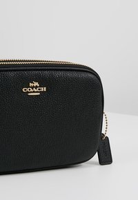 Musta nahkainen pussi, jossa on hiekkapintainen tekstuuri, kultainen vetoketju ja logo. Varustettu pienellä tagilla, joka on kiinnitetty ketjulla lisäyksikkönä.